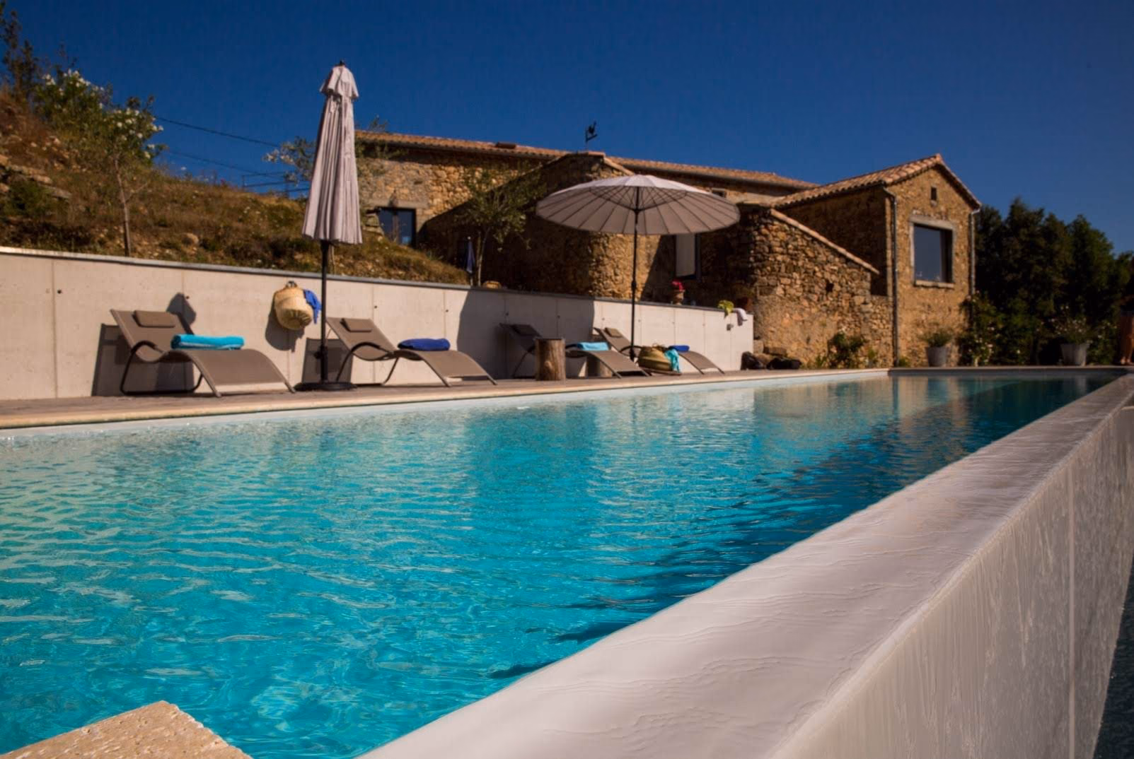 Mur de débordement blanc d'une piscine en béton, terrasse et maison en pierre en surplomb de la vallée