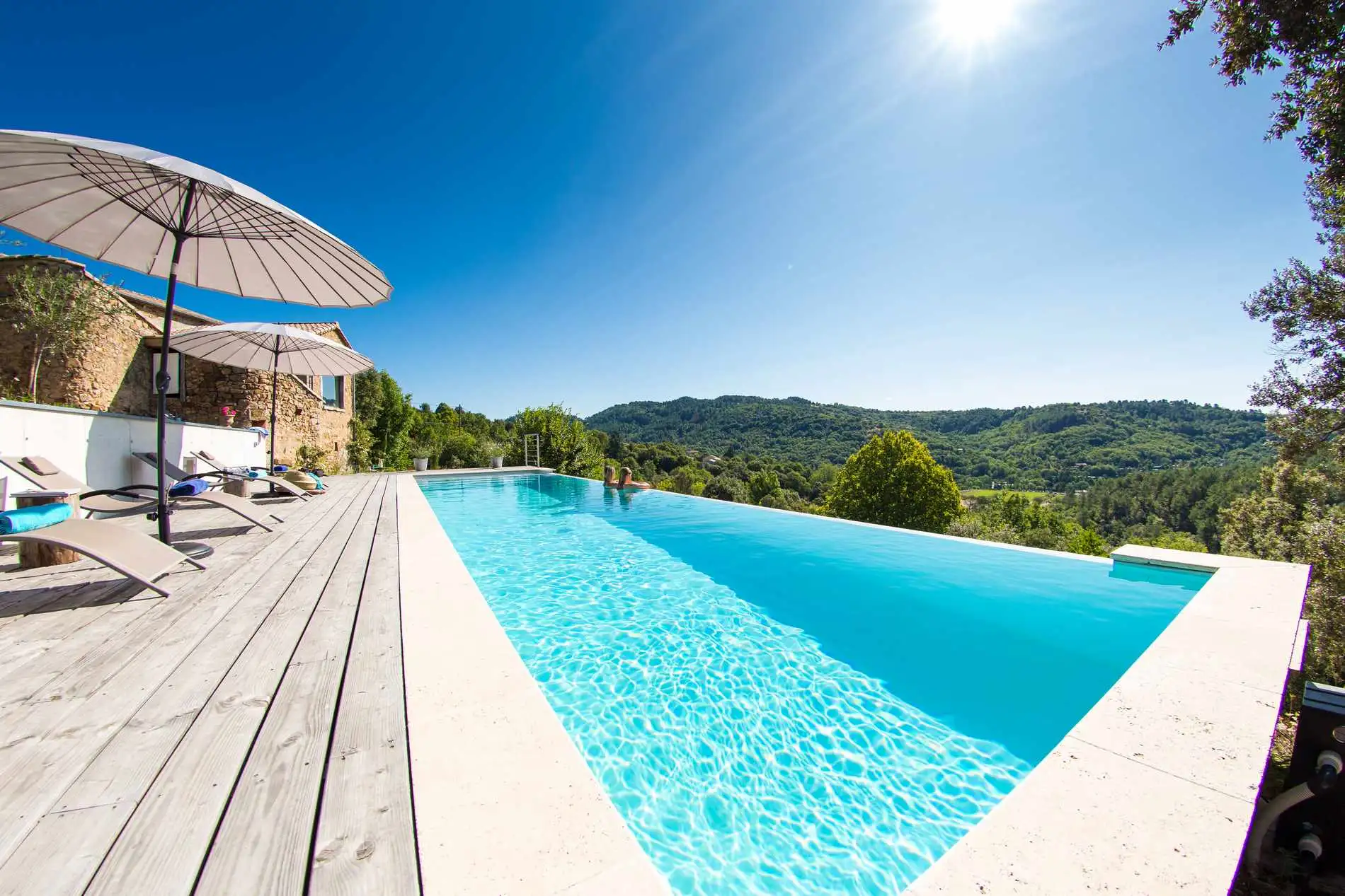 Piscine à débordement en béton armé surplombant une vallée ardéchoise, terrasse en bois et parasols