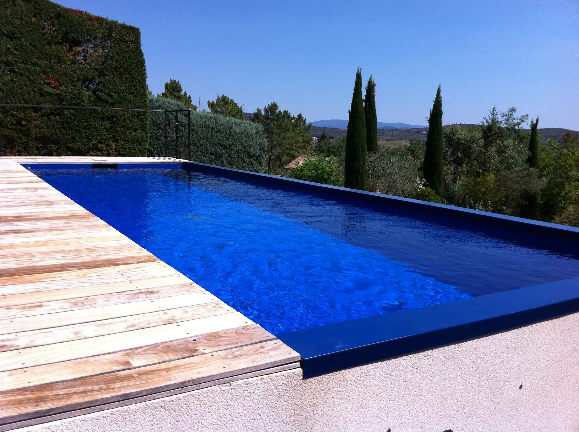 Piscine au revêtement bleu nuit et terrasse en bois face aux cyprès, en sud Ardèche