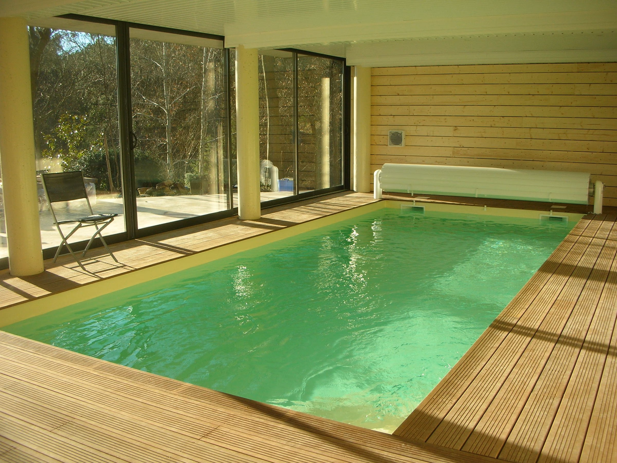 Piscine intérieure sous un volume vitré ouvert sur le jardin, plage en bois et volet hors-sol